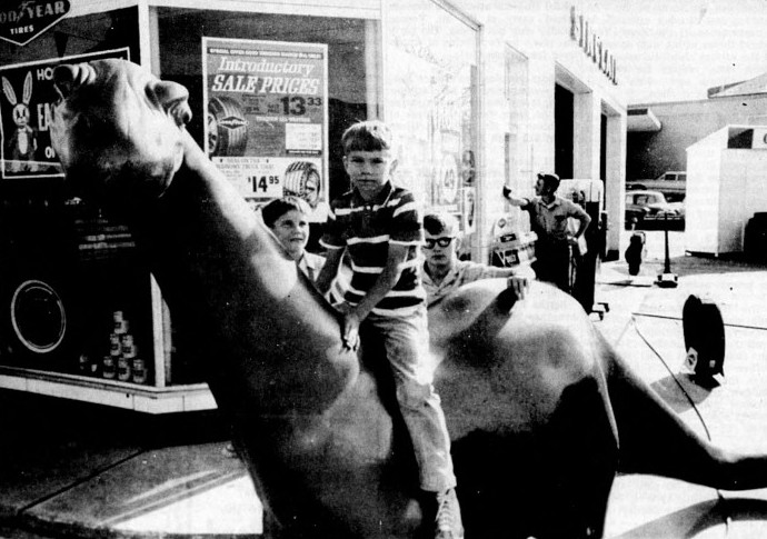 A boy sits on a dinosaur statue while two other boys look on.