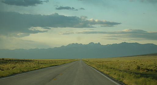 A road-level view of Los Caminos Antiguos Byway cutting through a flat valley towards the jagged Sangre de Cristo Mountains illuminated by the sunset.