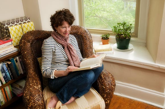 Author Paula McLain sitting in a wicker high back chare with her feet up and under her, sitting in front of a bay window. She is wearing jeans, a blue and white striped t-shirt and a mauve scarf. She is reading.