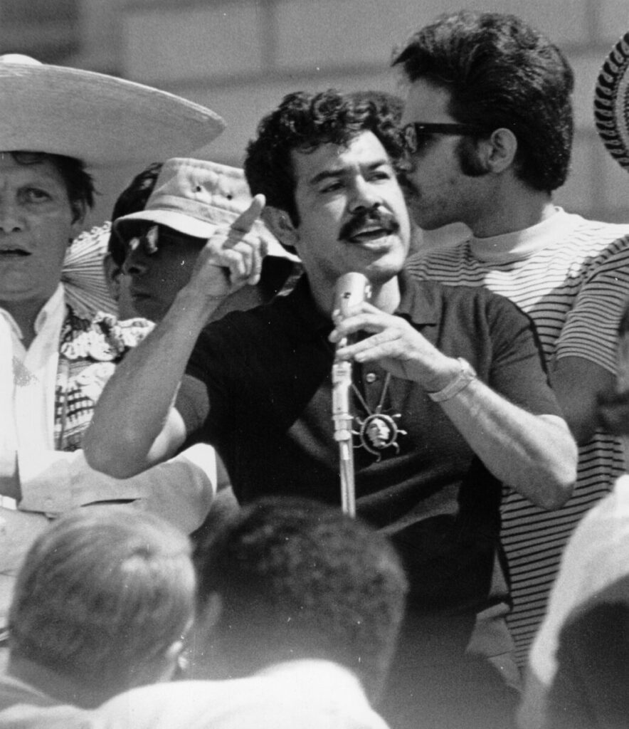 Photograph of Mexican American Rodolpho "Corky" Gonzales, Mexican American activist and founder of the Crusade for Justice, addresses a crowd in probably Denver, Colorado. He holds a microphone and wears a pendant. A man in a large straw sombrero stands nearby.