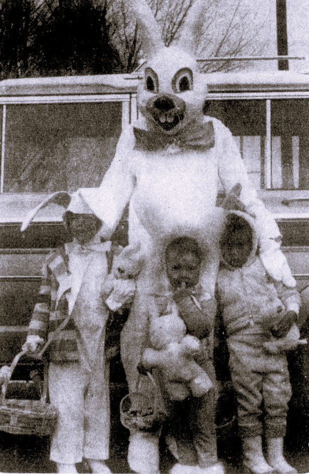 Three children wearing bunny ears stand beside the Easter Bunny.