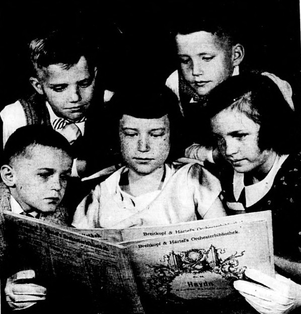 Five children wearing formal clothes look at a musical score.