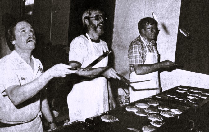 Three men flip pancakes at a large griddle.