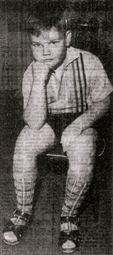 A boy wearing shorts, knee socks, and sandals poses in a chair.