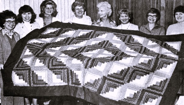Eight women hold up a quilt they made.