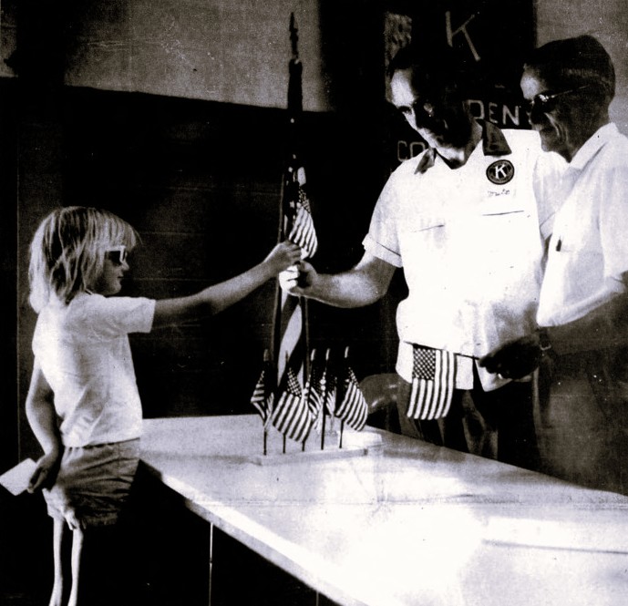 Two men give an American flag to a young girl wearing sunglasses.