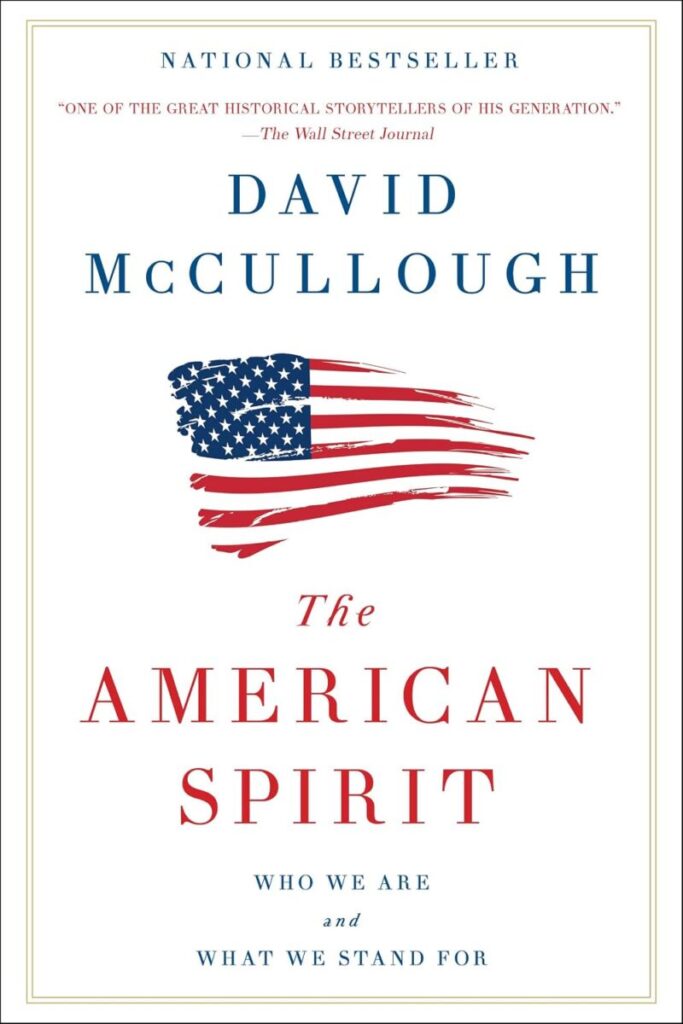Book cover for The American Spirit: Who We Are and What We Stand For by David McCullough. The cover features a clean, minimalist white background enclosed by a thin, elegant double border. At the top, the words "NATIONAL BESTSELLER" are printed, followed by a review quote from The Wall Street Journal. The author's name, "DAVID McCULLOUGH," is written in a classic blue serif font. In the center is a stylized illustration of the American flag, designed to look like a dynamic, horizontal brushstroke with tattered edges. Below the flag, the book's title is displayed in a sophisticated red serif font, with the subtitle printed at the very bottom in smaller, spaced-out gray text.