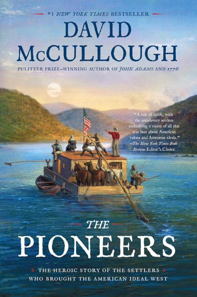 Book cover for The Pioneers: The Heroic Story of the Settlers Who Brought the American Ideal West by David McCullough. The top of the cover features the author’s name in large, elegant blue font, noting him as a "#1 New York Times Bestseller" and "Pulitzer Prize-winning author." The central illustration is a historic painting depicting a group of 18th-century settlers, including men, women, and horses, traveling down a wide river on a wooden flatboat. An American flag flies from a mast on the boat, and a man stands at the rear steering with a long sweep oar, pointing toward the forested hills and sunlit sky in the background. The book's title, "THE PIONEERS," is printed across the bottom in large white block letters against the blue river water.