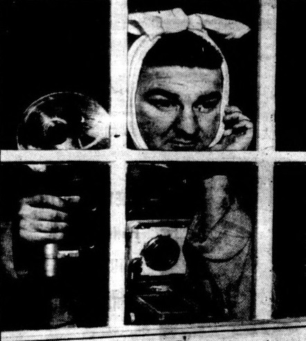 A man wearing a mumps head wrap stands behind a window holding a camera.