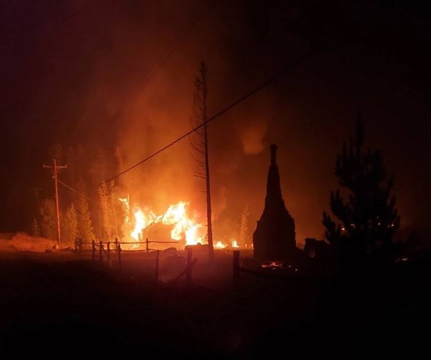 Night time view of a burning home. In the foreground is a lone chimney. Burned trees and power lines cut across the image.