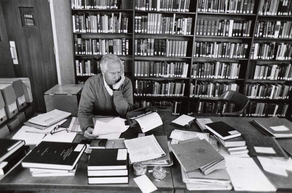 A black and white historical photograph of author David McCullough working at a large desk filled with research materials. McCullough, a white man with gray hair wearing a sweater over a collared shirt, sits behind the desk resting his chin in his left hand, looking down intently at a document he holds in his right hand. In the center of the desk sits a vintage manual typewriter with a sheet of paper fed through it. The expansive desk is cluttered with numerous stacks of books, open folders, loose papers, a pair of eyeglasses, and small containers holding paper clips. Spanning the entire wall behind him are massive, floor-to-ceiling wooden bookshelves densely packed with rows of books.
