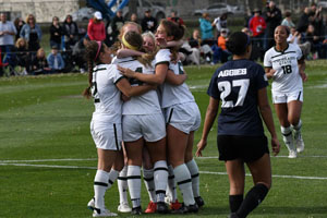 A group of 5 soccer players celebrate with a group hug on the field as one of their teammates runs toward them from the right. A player from the other teams walks away from the celebration. Spectators sit on the sidelines in the background.