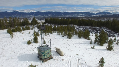 An aerial photograph of a cloud seeding generator sitting on a snowy hill surrounded by small evergreen trees. A mountain range is visible in the distance. The generator is comprised of a large, camouflaged metal box with a long skinny chimney ending in metal cannister that contains a flame used to disperse particles into the air.
