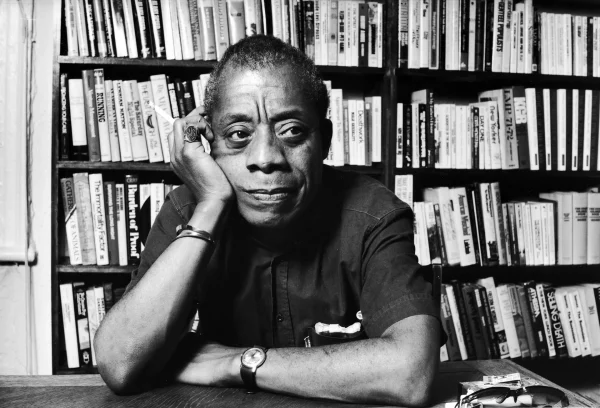 Black and white photo of James Baldwin sitting in front of a bookshelf, holding a cigarette in his right hand. His right palm is holding his head with his elbow on the desk in front of him and he is looking left of the camera