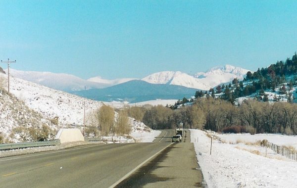 Cars drive down a paved portion of the Colorado River Headwaters Scenic and Historic Byway towards high mountain peaks covered in snow.