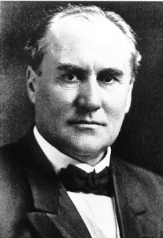 A black and white photographic headshot of Governor Buchtel, who has a serious facial expression and is wearing a black jacket, white shirt, and a black bowtie. 
