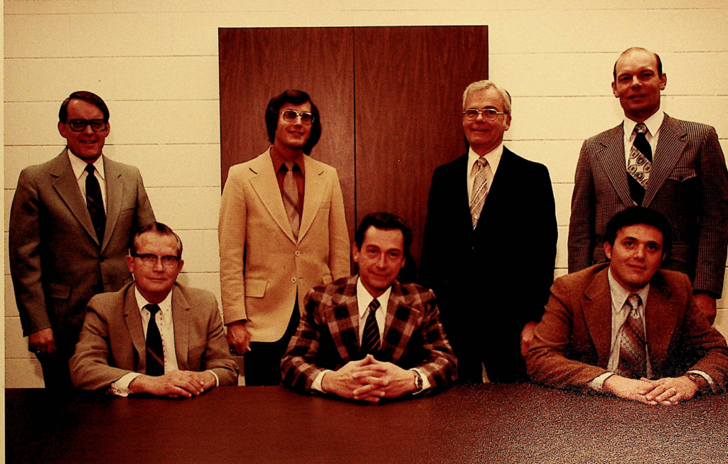 Group of seven men in suits and ties; three man sit at a table, their hands folded in front of them; four others stand behind them