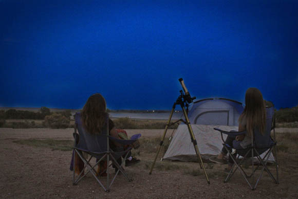 Two people sit facing away from the camera on camp chairs in a plains landscape at dusk, looking up at the night sky with a telescope pointing towards the sky at the center of the image