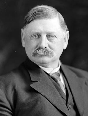 Black and white photographic headshot of Governor John Shafroth around age 55. In the photo, he wears a black suit jacket, a white collared shirt., a black vest, and a necktie. He sports a prolific mustache.