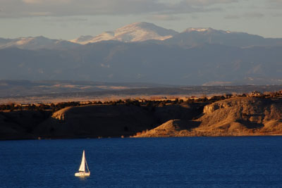 A landscape photograph showing Lake Pueblo at sunset. The dark blue lake is in the foreground. A white sailboat is floating on the lake in the lower left corner of the image. The sailboat is dwarfed by the large lake and the surrounding landscape of buttes and prairie plants. Snow-covered mountains tower in the background. 