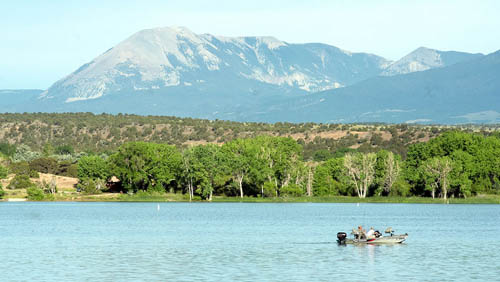 A large light blue lake in the foreground, with a fishing boat carrying two men on the right side of the photograph. Green, leafy trees line the banks of the lake and the Spanish Peaks mountains tower in the background. The mountains are covered with greenery until the treeline ends, with grey rocks exposed at the top of the mountains.