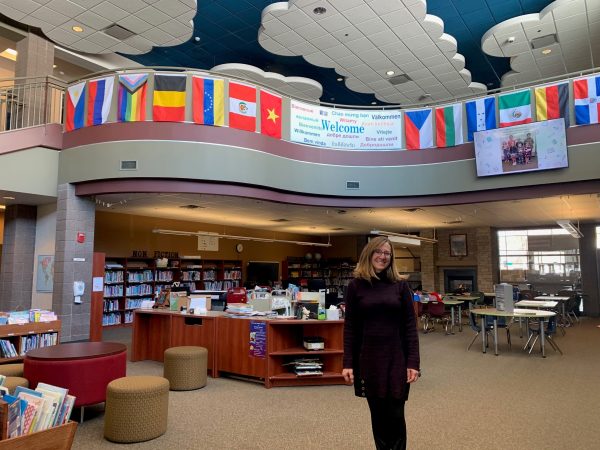 Soda Creek Elementary teacher librarian Libby Creamer in front of the library's desk, with a 'welcome' banner and flags on the balcony above