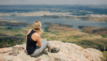 A photograph of a woman sitting with her back to the camera. She sits on a rocky outcropping looking out onto the plains below, featuring a large reservoir.