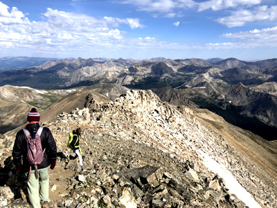 Two people walk down a rocky ridge high above treeline. Many other tall mountain peaks are visible in the distance.