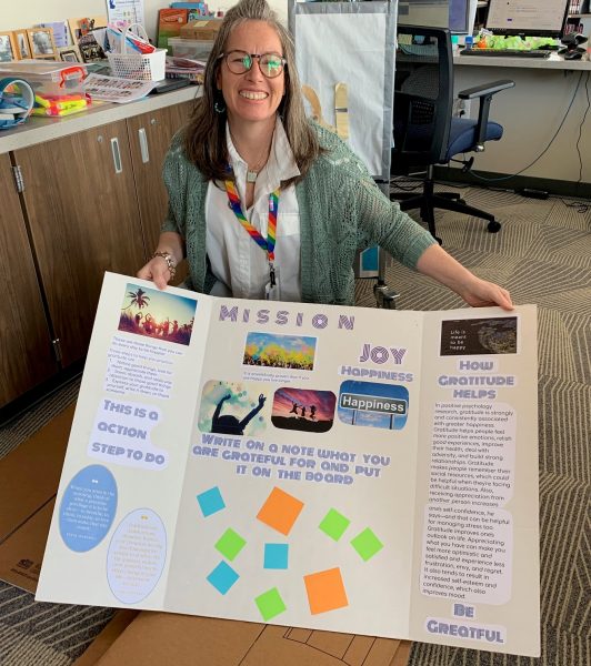 Photo of teacher librarian Nicole DeCrette with a trifold display created by students for their Mission Joy station about gratitude,