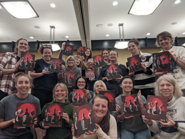 Group of adults posing and holding up the same painting of a dark wine glass and wine bottle before an eerie red backdrop 