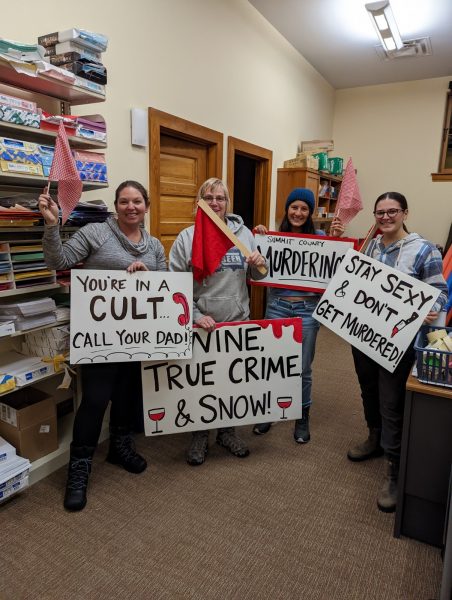 group of smiling women holding up quotes from a popular true crime podcast on signs including youre in a cult call your dad and stay sexy and don't get murdered