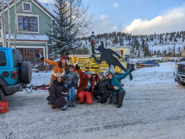 group of people posting in front of a sled decorated with crime scene tape and a chalk outline of a troll made to look like a dead body. it is snowy and they're smiling 