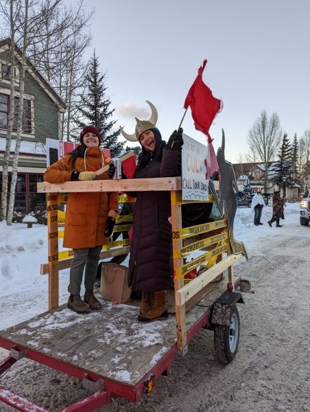 two smiling women on a wagon decorated with crime scene tape. one wears a viking horn hat and both are smiling. it is snowy