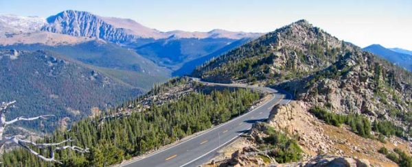 An aerial view of a two-lane road cutting along a mountain ridge right about at tree line, with a high mountainous landscape visible in the background.