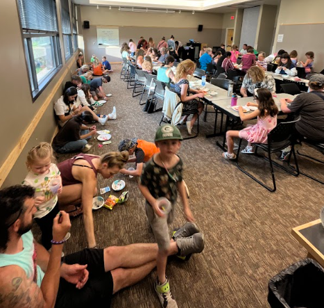 a room full of people of all ages sitting at tables and on the floor with beads and strings