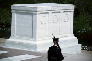 soldier with rifle on shoulder in front of white marble sarcophagus. 