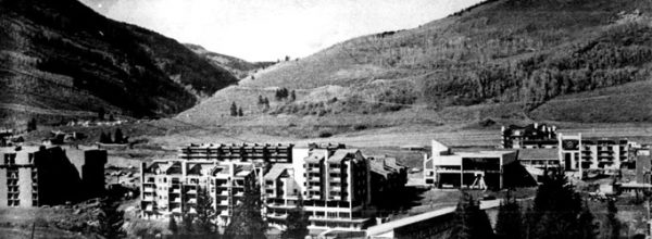 A black and white photograph, featuring condo buildings in the foreground and the snow-free hills of Vail's ski resort in the background.