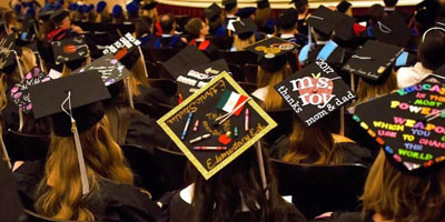 A view of about twenty students facing away from the camera sitting in rows of chairs during a commencement ceremony. The students are wearing black commencement robes. The angle of the photo shows the tops of their decorated graduation caps.