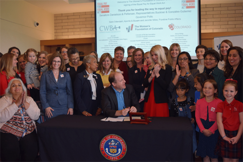 Governor Polis sits at a desk at the center of the photo, surrounded by a crowd of people as he prepares to sign paperwork laid out on the desk.