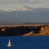 A landscape photograph showing Lake Pueblo at sunset. The dark blue lake is in the foreground. A white sailboat is floating on the lake in the lower left corner of the image. The sailboat is dwarfed by the large lake and the surrounding landscape of buttes and prairie plants. Snow-covered mountains tower in the background.