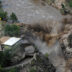 An aerial photograph of a flooded river thundering down a small waterfall directly to the right of a house that sits on the right of a highway. The river water is brown with sediment and is creeping up the sides of the canyon walls to the left and the house and road to the right. The surrounding trees have remained intact and there is little debris in the river.