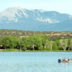 A large light blue lake in the foreground, with a fishing boat carrying two men on the right side of the photograph. Green, leafy trees line the banks of the lake and the Spanish Peaks mountains tower in the background. The mountains are covered with greenery until the treeline ends, with grey rocks exposed at the top of the mountains.
