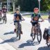 A teacher leads a line of school-age kids riding bikes, all wearing matching t-shirts with their school logo.